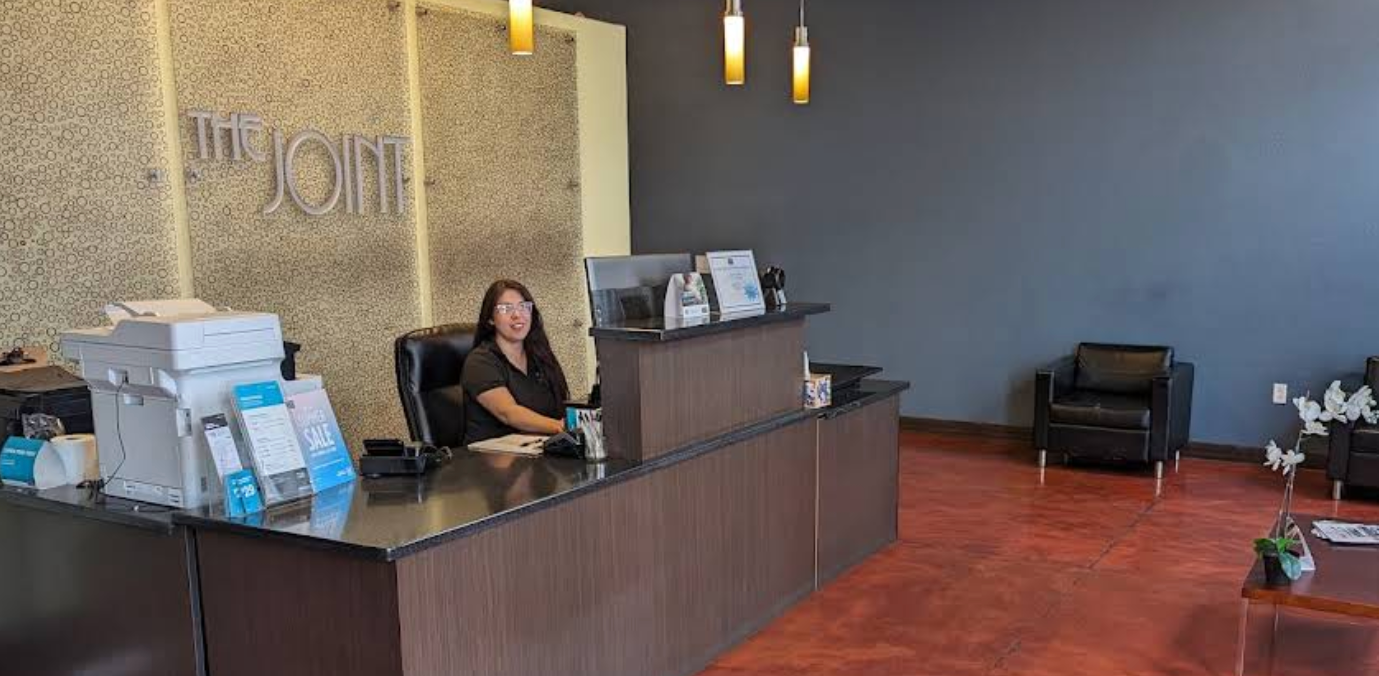  A female receptionist smiles from behind a dark wood front desk at a clinic reception area. The background features a textured wall with "THE JOINT" signage on the left, modern hanging pendant lights, a dark grey accent wall, and a waiting area with two black leather armchairs and a small coffee table on a polished reddish-brown concrete floor.
