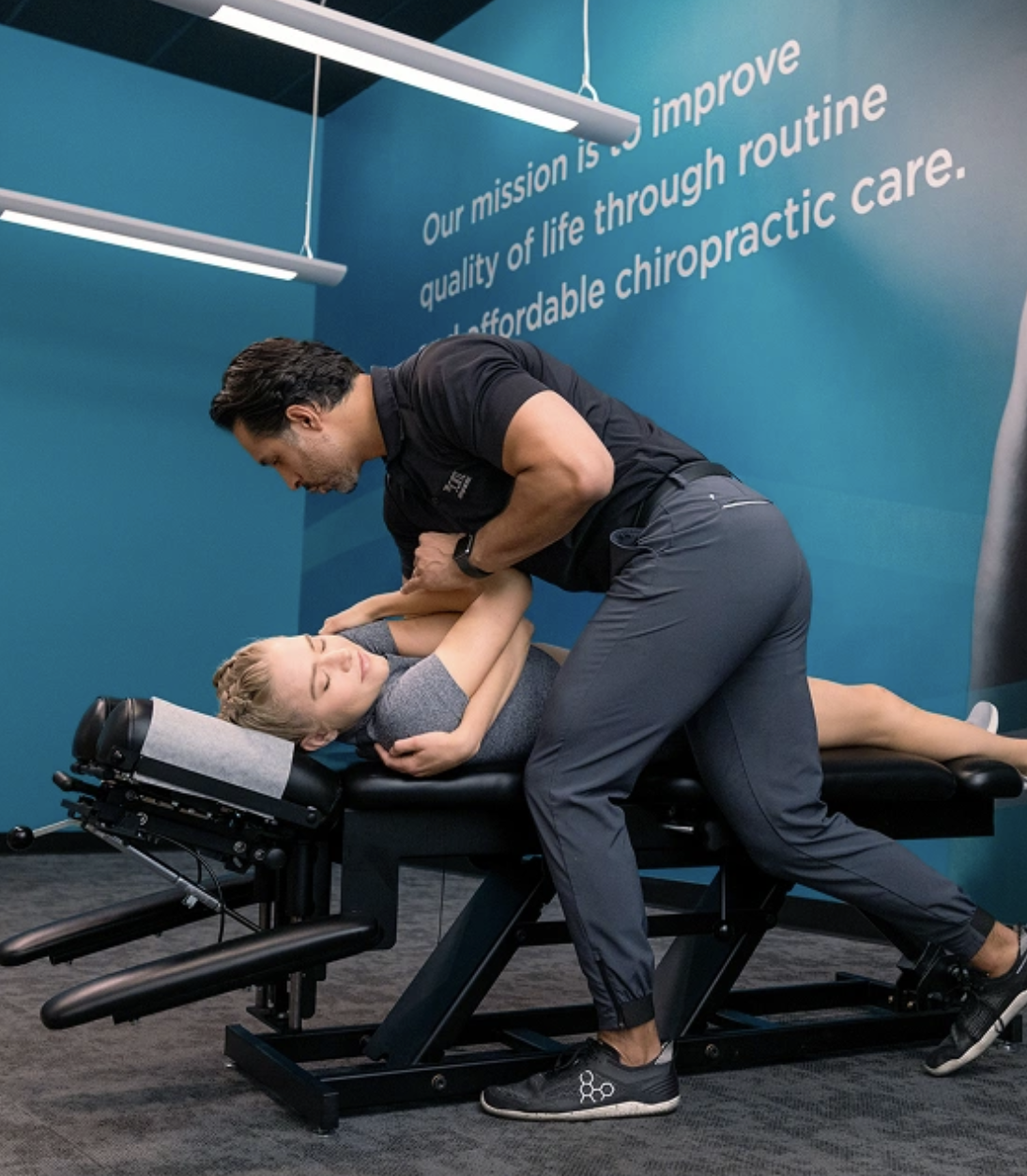 A male chiropractor in a black polo shirt and grey pants performs a manual spinal adjustment on a female patient lying on her side on a black chiropractic table. The setting is a clinic with bright overhead lights and a teal accent wall featuring text that reads, "Our mission is to improve quality of life through routine and affordable chiropractic care."