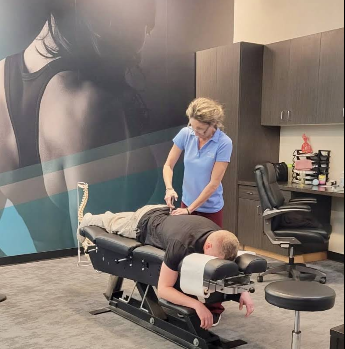 A female chiropractor in a light blue polo shirt uses a handheld activator adjusting instrument on a male patient's lower back as he lies face down on a black chiropractic adjustment table in a clinic office.