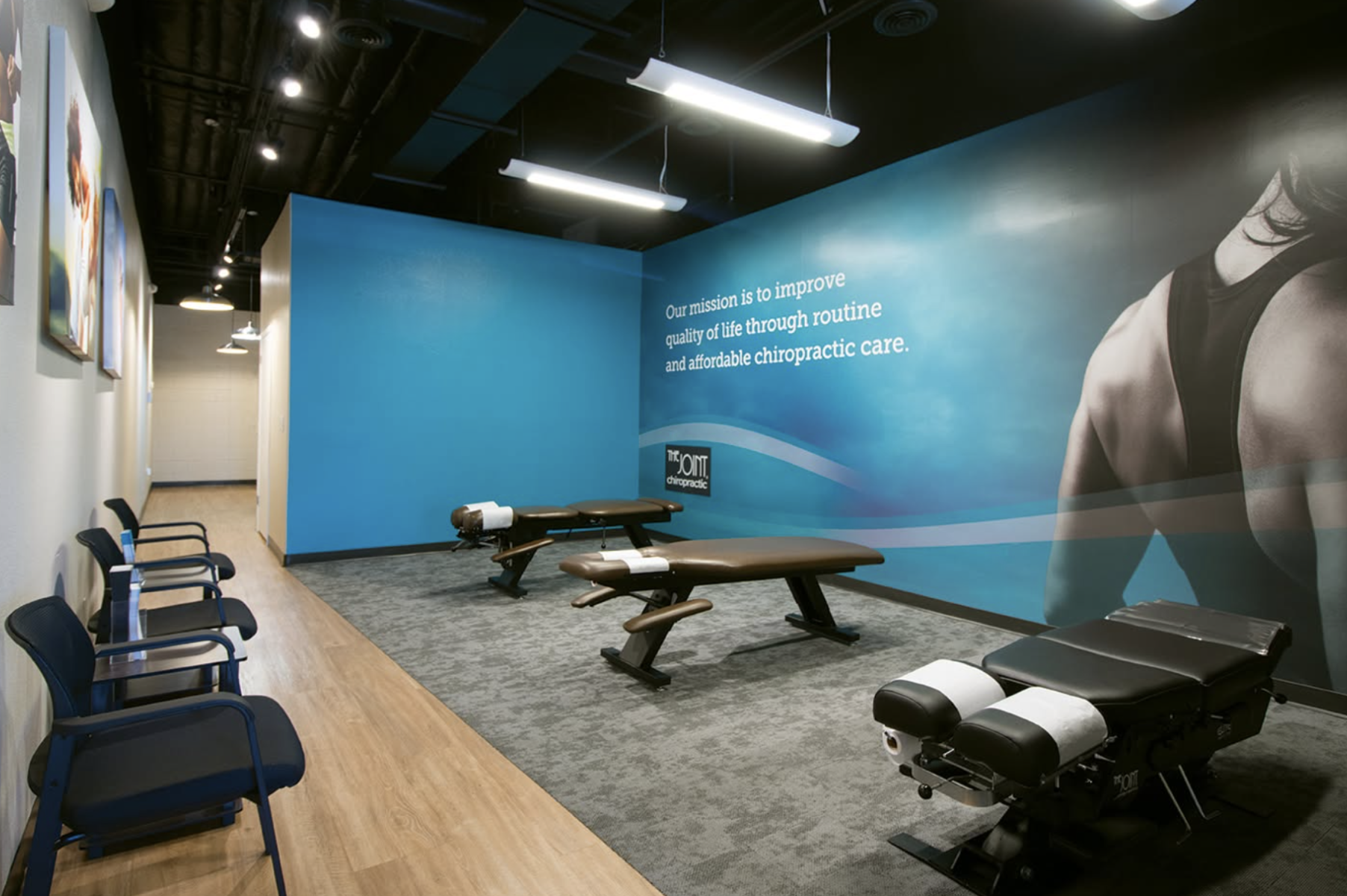 Modern treatment room at The Joint Chiropractic clinic featuring multiple adjustment tables, blue feature walls with mission signage, and seating for patients.