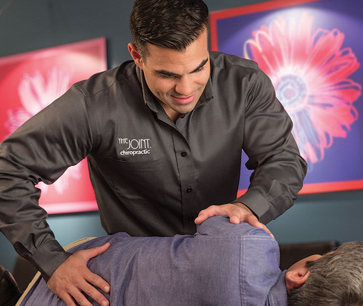 A licensed chiropractor wearing a grey button-down shirt with the "The Joint Chiropractic" logo performing a side-posture lower back adjustment on a male patient in a wide, landscape-ratio clinical setting.