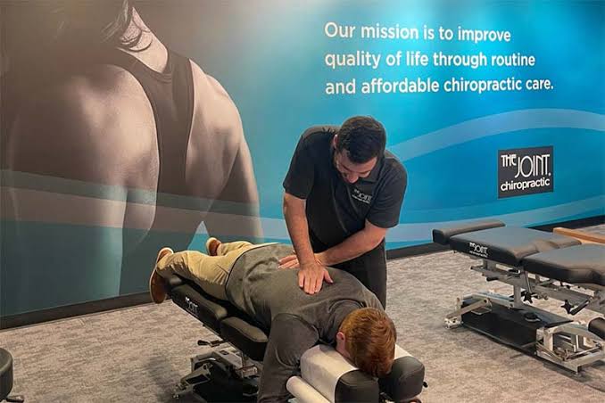 A licensed doctor of chiropractic at The Joint Chiropractic performing a manual spinal adjustment on a male patient lying face down on a professional adjustments table, with the brand's blue mission wall visible in the background.