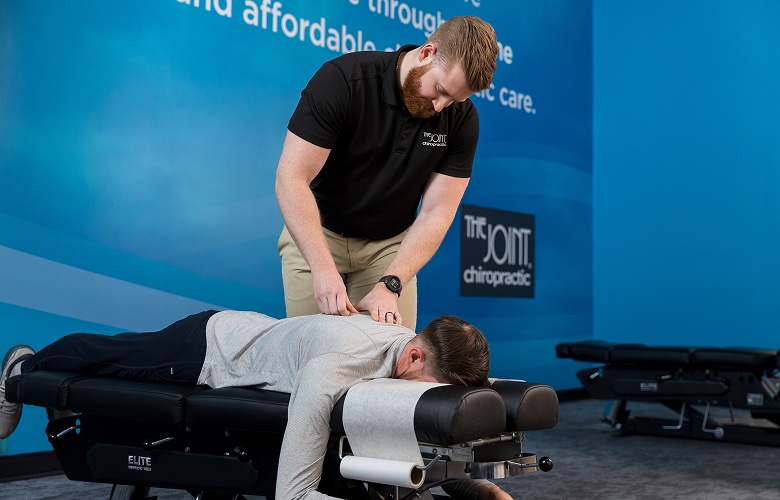 A licensed chiropractor with a red beard, wearing a black polo shirt with "The Joint Chiropractic" logo, examines a male patient's upper back and spine as the patient lies face down on a black Elite chiropractic adjustment table against a blue brand wall.
