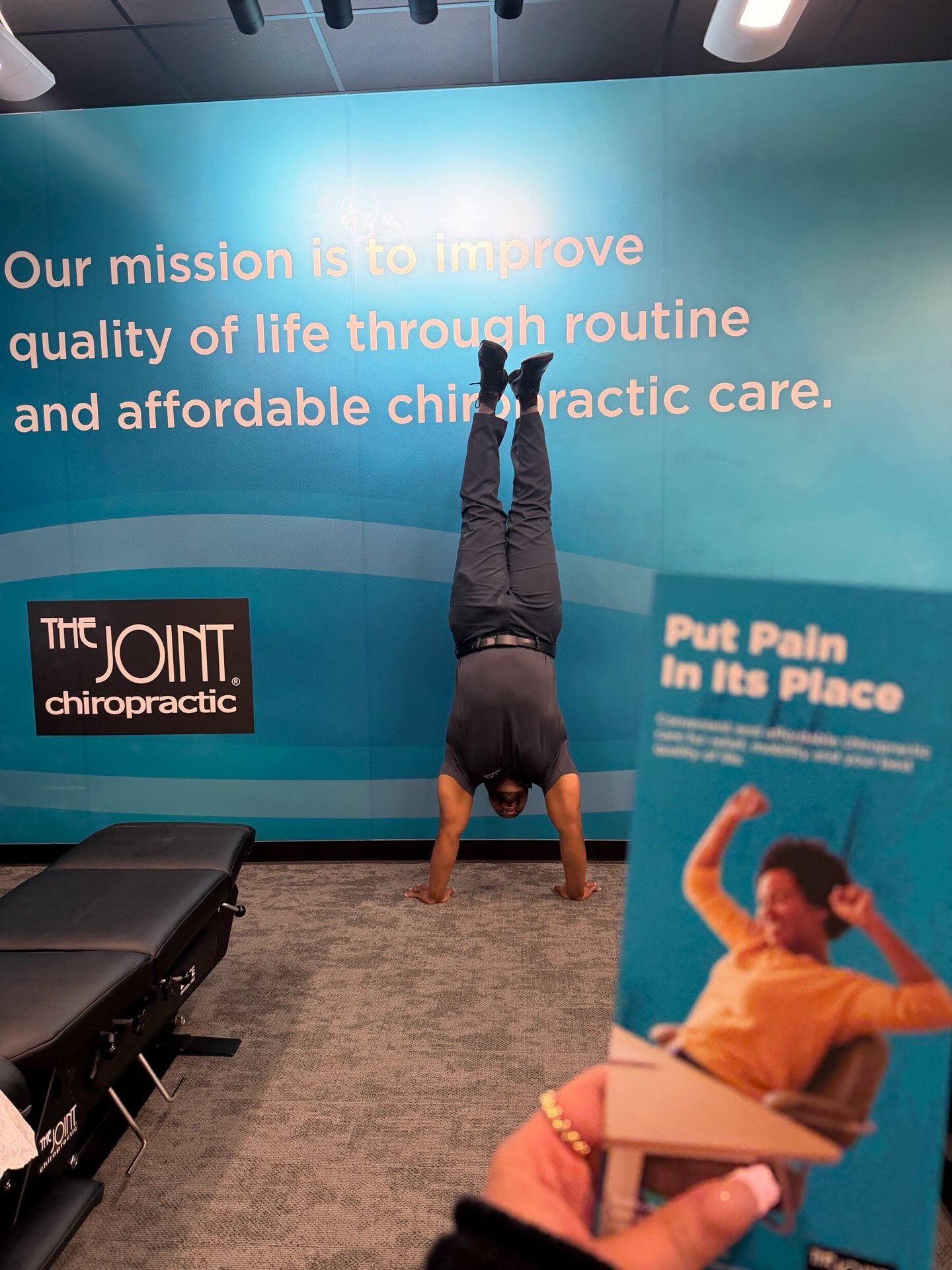 A Doctor of Chiropractic demonstrating full-body balance and mobility by doing a handstand inside The Joint Chiropractic clinic near a brand wall and promotional brochure.