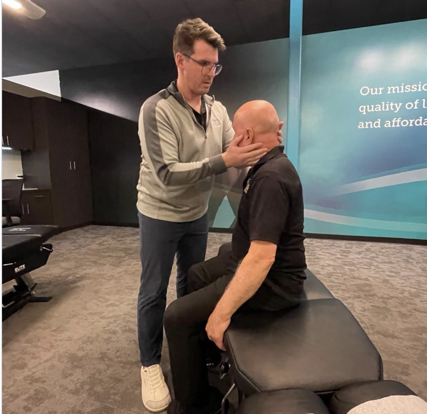 A Doctor of Chiropractic standing beside a seated male patient while gently evaluating his neck and cervical spine mobility inside a modern clinic setting.