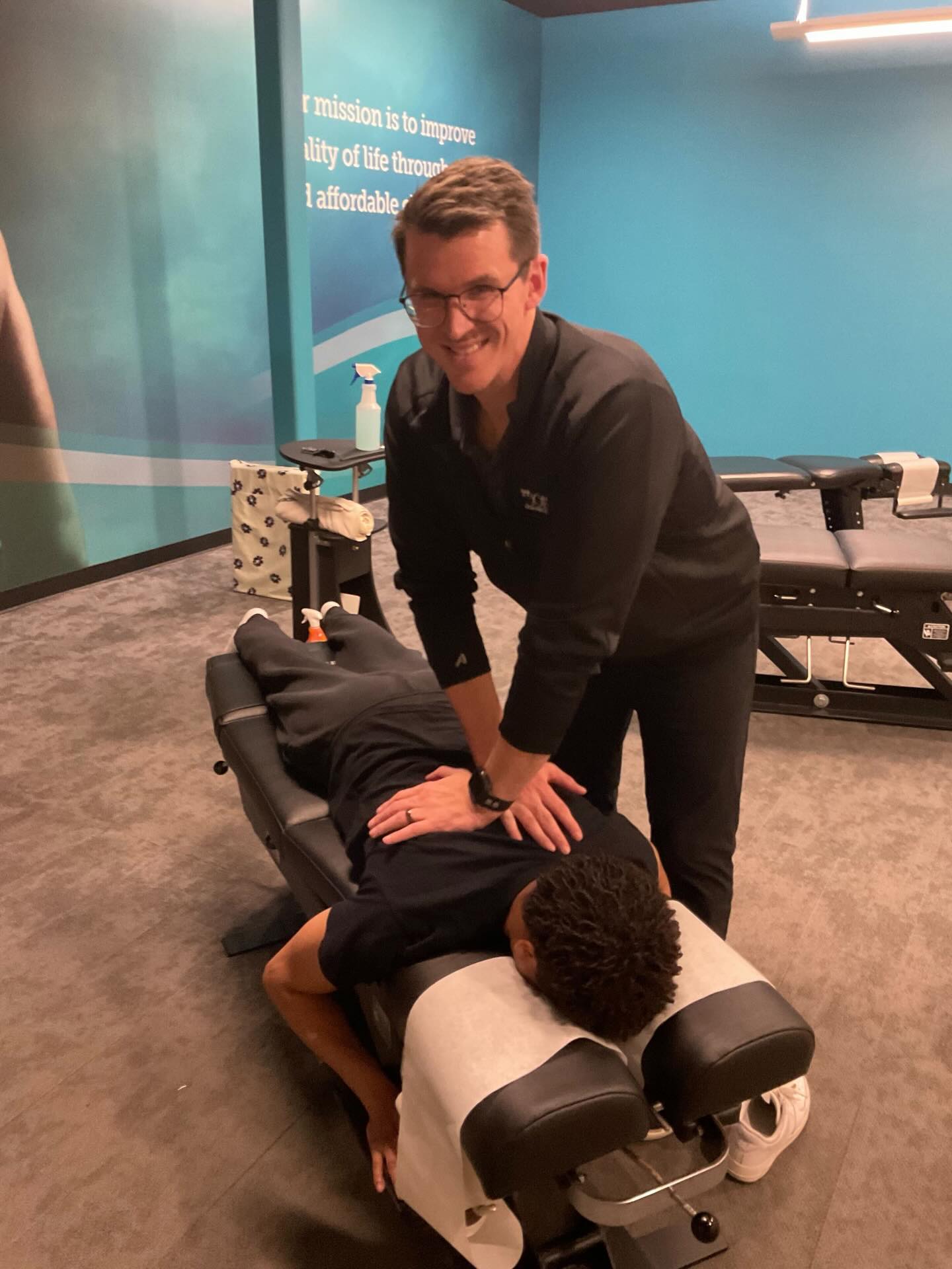 A smiling Doctor of Chiropractic performing a spinal adjustment on a male patient lying prone on an adjustment table at The Joint Chiropractic.